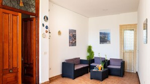 Een ruime woonkamer met een houten deur, twee donkere rieten banken, een klein bijzettafeltje en een potplant.