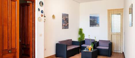 Uma sala ampla com porta de madeira, dois sofás de vime escuro, uma mesinha lateral e um vaso de planta.