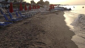 A beach with rows of blue and white sun loungers, red umbrellas, and a calm sea. - SimOtel Ermis (Kassandra)