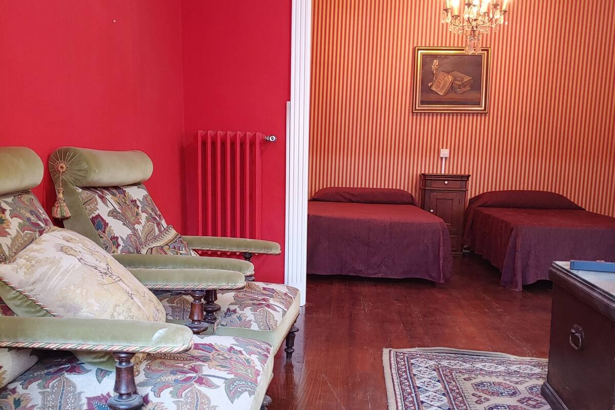Una habitación con pared roja, sofá estampado, suelo de madera, alfombra con diseño y cama con colcha roja.