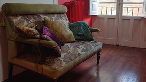 A room with a wooden floor, a green sofa with patterned cushions, a red radiator, and a white door with a view of the outside. - El Bordón de la Casa Batallón (Sarria)