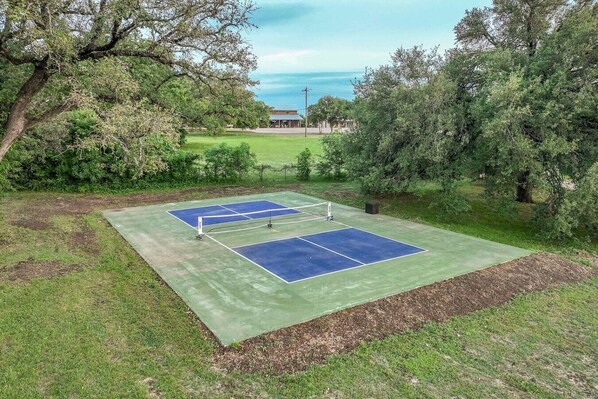 An outdoor tennis court with a net, surrounded by trees and grass.