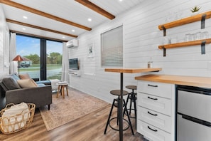 A compact kitchen with a built-in stove, wooden shelves, a small dining area with stools, and a large window offering a view of the outdoors.