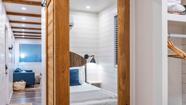 Une chambre à coucher comprenant un lit, une table de chevet, une lampe et donnant sur un couloir où se trouvent un canapé bleu et un panier.