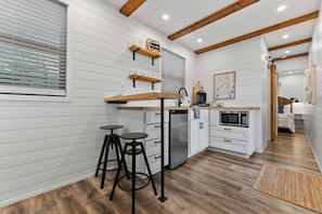 A compact kitchen with white paneling, wooden beams, a built-in microwave, and a small dining area with stools.
