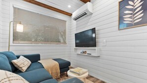 Un salon avec un canapé bleu, une petite table ronde, un téléviseur fixé au mur et une fenêtre avec des stores.
