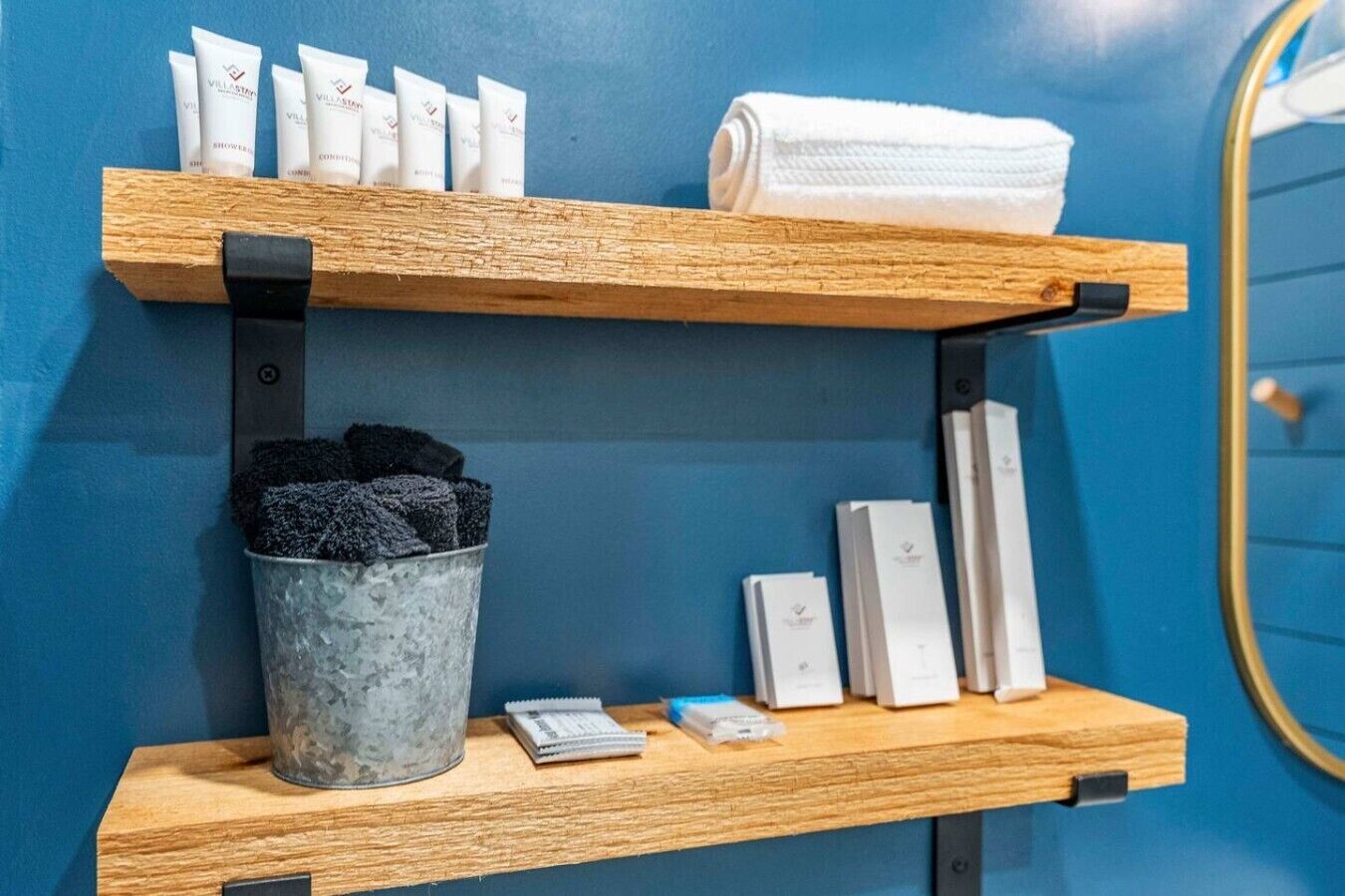 Un baño de hotel con estantes de madera que sostienen artículos de aseo, una toalla y un cubo con toallas negras.