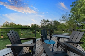 A wooden deck with Adirondack chairs and a small table, overlooking a grassy area and trees.