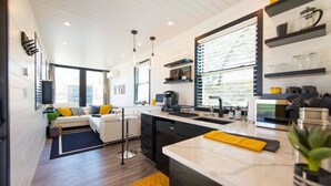 A modern kitchen with a central island, built-in appliances, and a large window providing natural light. - Tiny Container Home with Rooftop Deck (Waco)