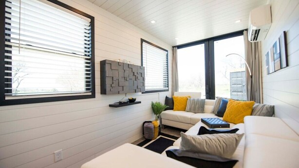 A modern living room with a sofa, a coffee table, and a wall-mounted TV. There are windows with blinds, a ceiling fan, and a wall-mounted air conditioner. - Tiny Container Home with Rooftop Deck (Waco)