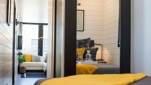 A modern bedroom with a bed, bedside table, and a view of a living area with a sofa and a plant. - Tiny Container Home with Rooftop Deck (Waco)