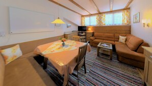 Ein gemütliches Zimmer mit Holzbalkendecke, einer Couch, einem Tisch mit floralem Tischläufer und einem Fernseher.