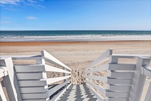 Una entrada a la playa con una escalera de madera blanca que baja a la arena.