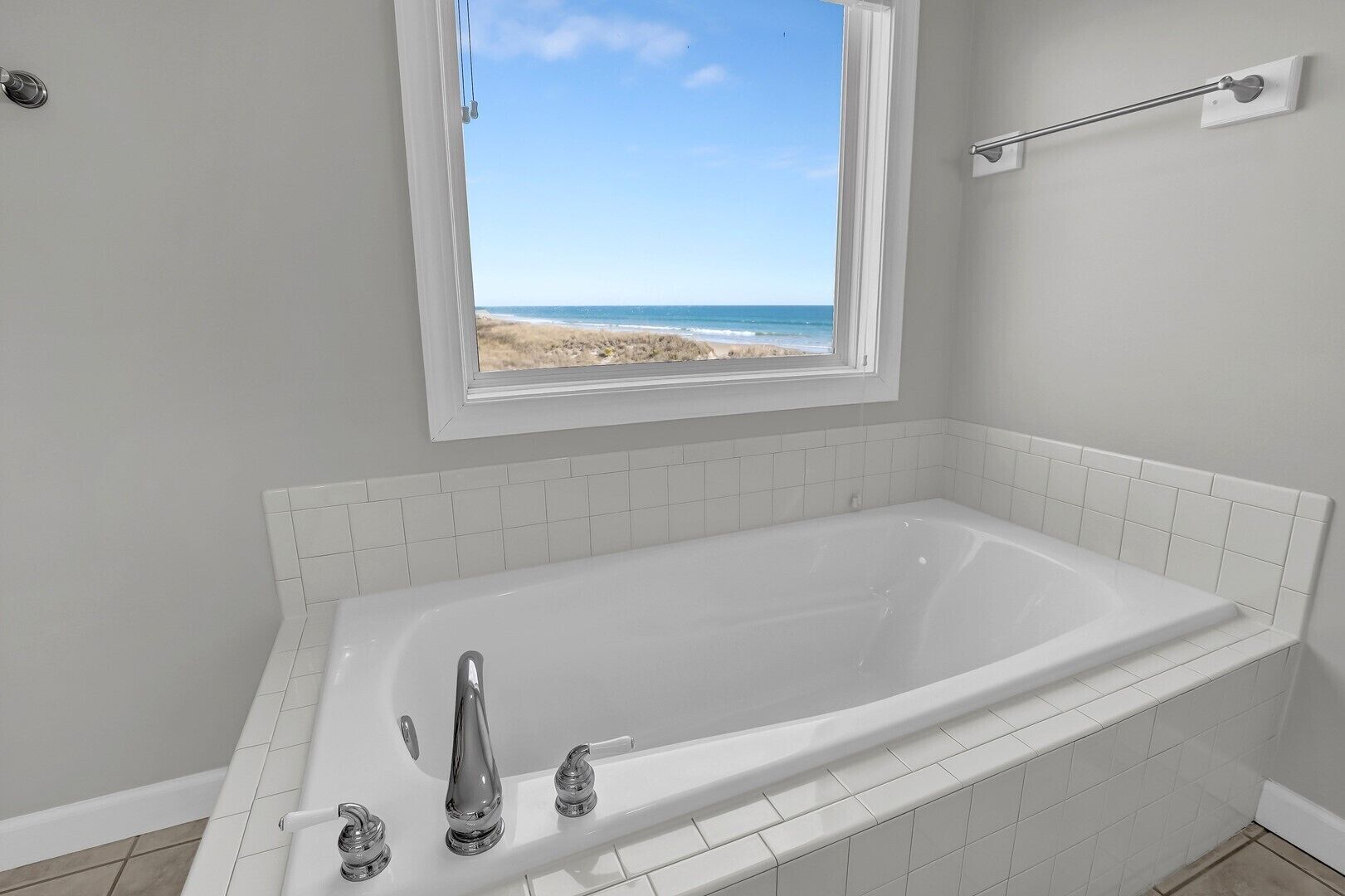 Et badeværelse med et stort badekar, et vindue med udsigt til stranden og hvide flisemure.