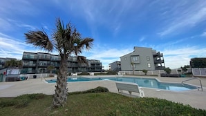 A pool area with a palm tree, a bench, and modern buildings in the background. - Amazing views of the ocean, community pool, private covered deck (North Topsail Beach)