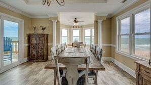 A dining room with a large wooden table, white chairs, and a chandelier. There are large windows with a view of the beach. - 8 Bedroom Direct Oceanfront Home, Private Pool, Media Room, Private Beach Access, 2025 Updates (North Topsail Beach)