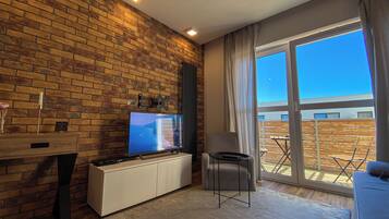 A modern living room with a brick wall, a flat-screen TV, a white entertainment unit, a small table with a remote, and a large sliding glass door leading to an outdoor area.