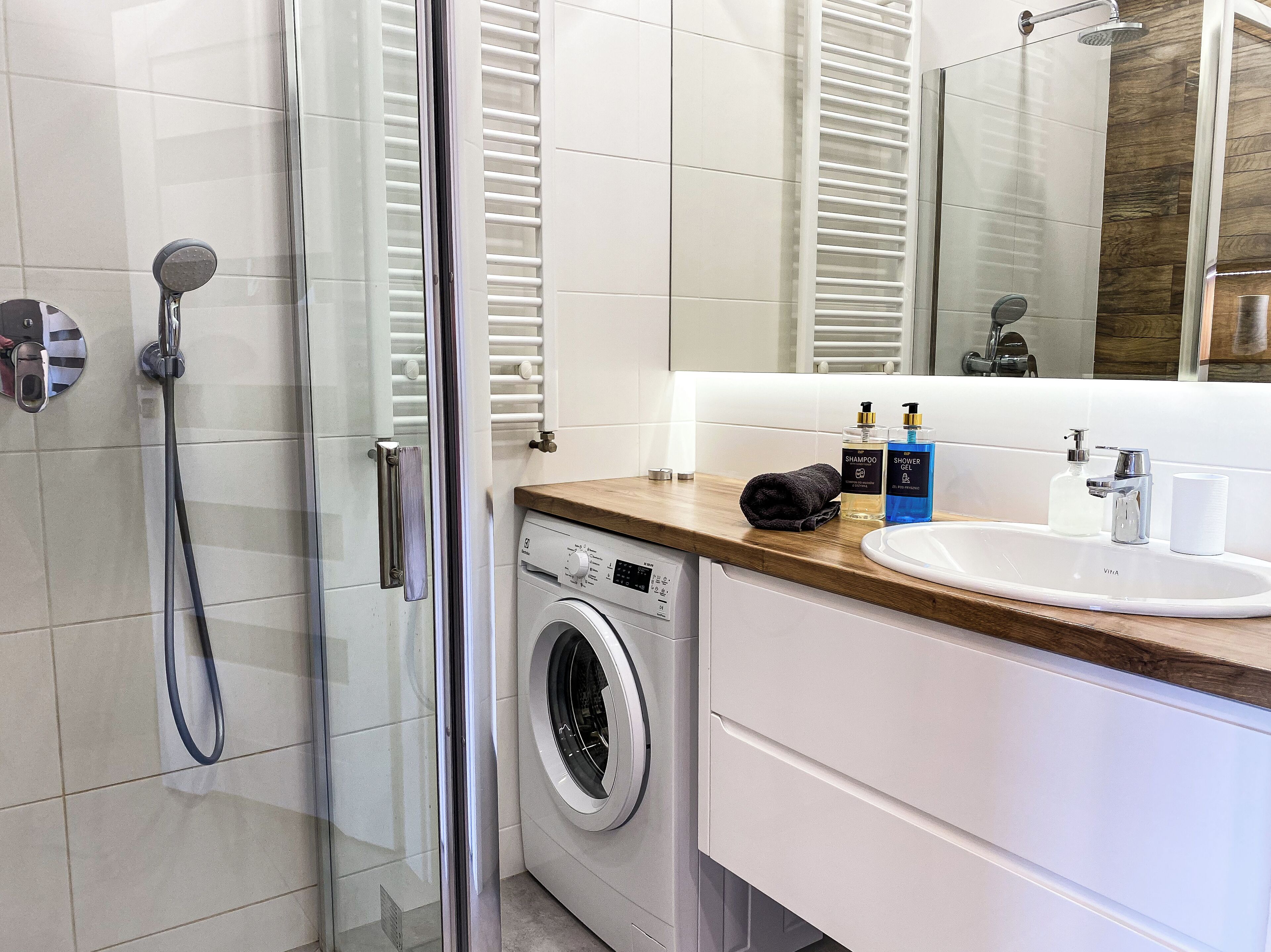 Ein modernes Badezimmer mit Duschkabine aus Glas, integrierter Waschmaschine und Waschtisch mit Spüle und Pflegeprodukten.