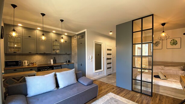 A modern kitchen with built-in cabinets, a dining area with a sofa, and a bedroom with a large window. - BP Apartment I (Torun)