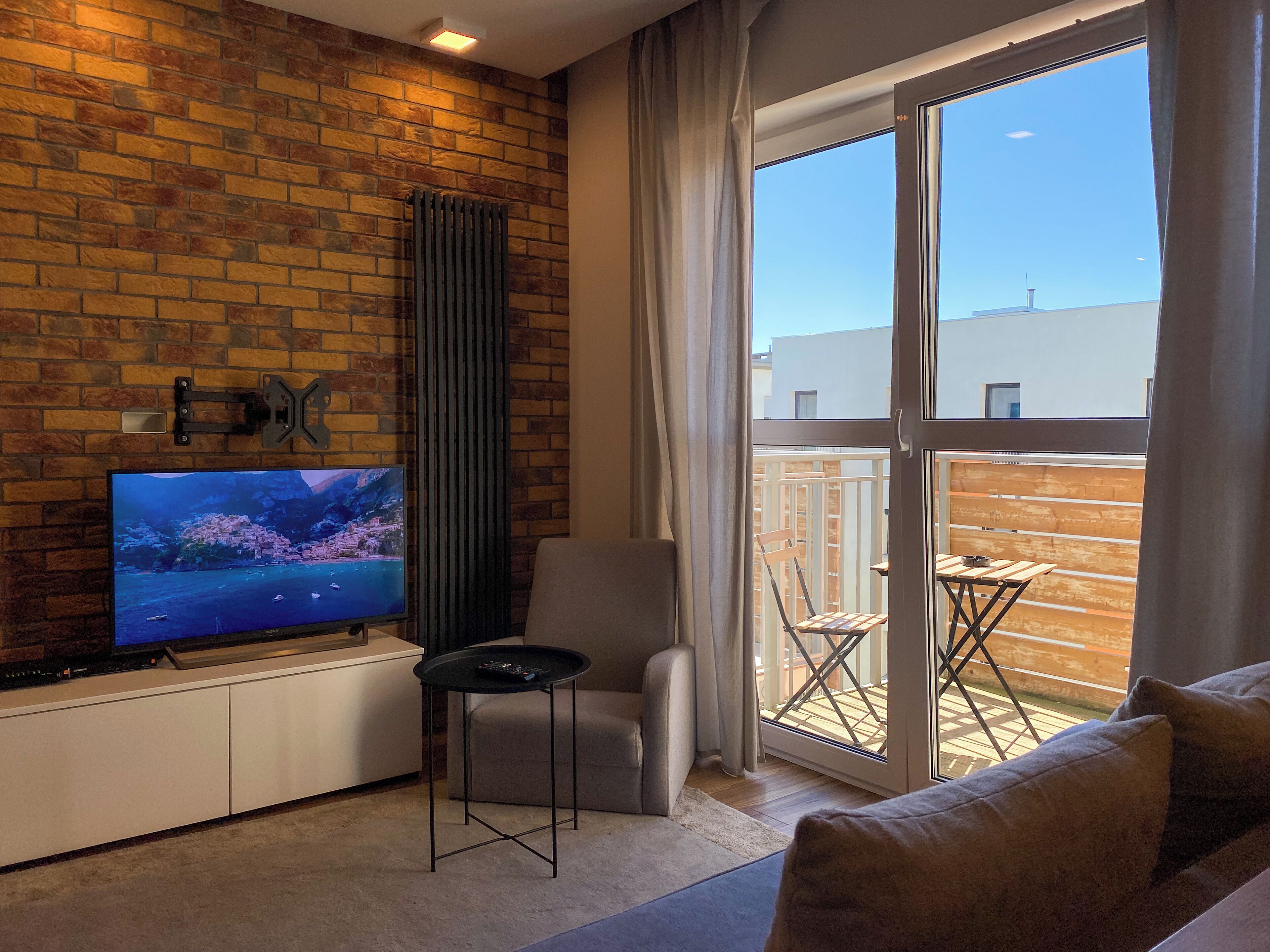 A modern living room with a brick wall, a flat-screen TV, a grey armchair, a small round table, and a large window with a view of the outside.