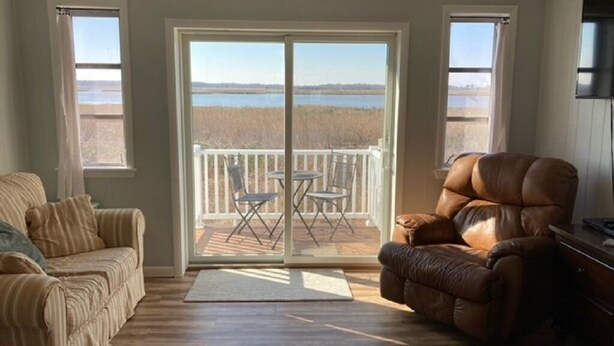 Un salón con un gran ventanal corredera que da a un balcón con vistas a un cuerpo de agua y árboles. Hay un sillón de cuero marrón y un sofá a rayas.
