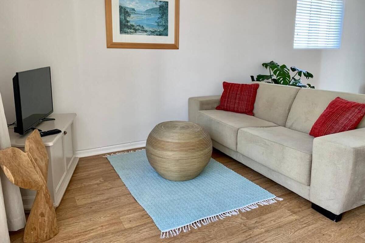Ett vardagsrum med en beige soffa, en rund pall, en inramad tavla på väggen, en tv på ett tv-bord och ett fönster med persienner.