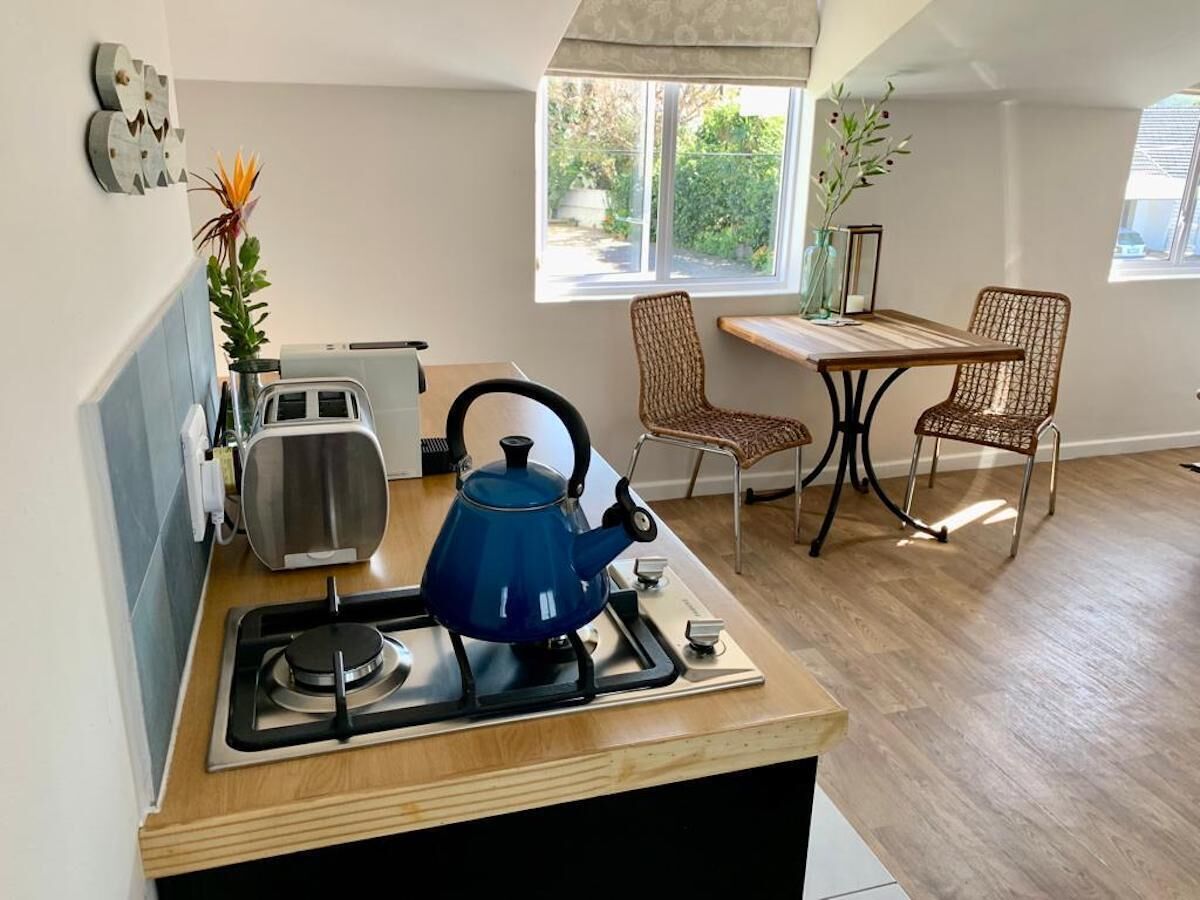 A compact kitchen with a gas stove, toaster, and blue kettle. There are two wicker chairs and a small table with a vase of flowers.