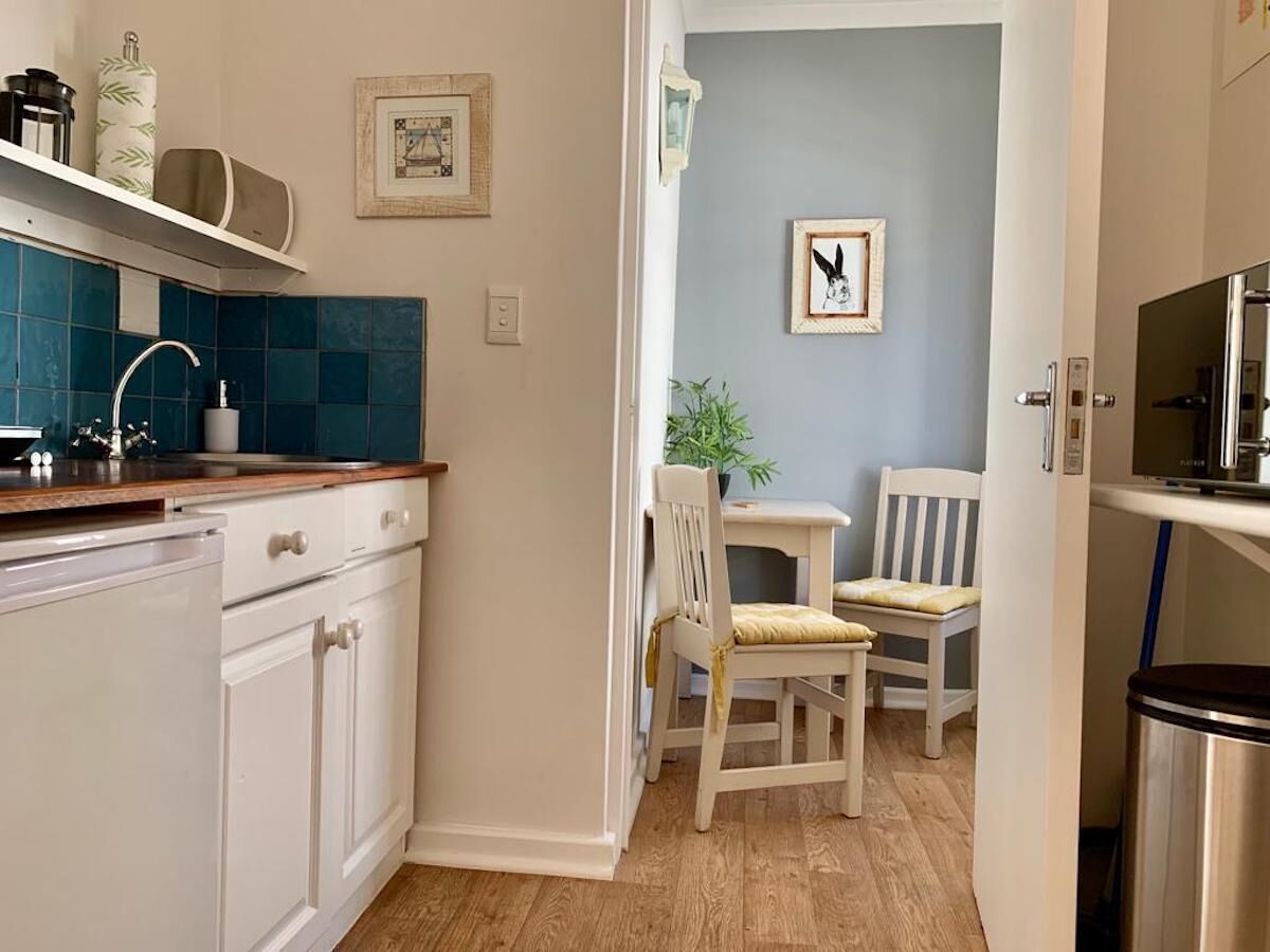 Une cuisine avec des armoires blanches, un pare-feu bleu et un plan de travail en bois. On y trouve une chaise blanche avec un coussin, une petite table et un tableau encadré accroché au mur.
