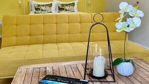 A yellow sofa with a cushion, a glass candle holder, a book titled "Cape Whale Coast," and white orchids on a wooden table. - Anchor's Rest Hermanus (Hermanus)
