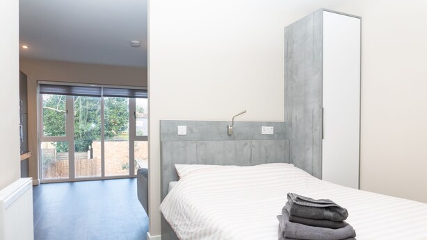 A bedroom with a bed, a grey headboard, a white wall, and a view of a garden through a sliding door. - Magna House (Egham)