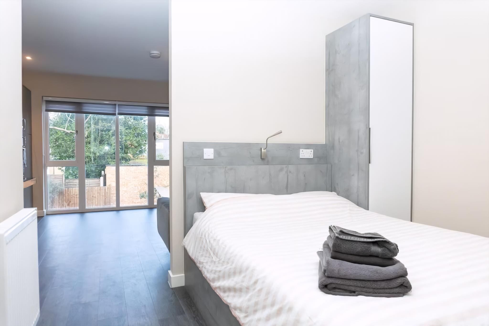 A bedroom with a bed, a grey headboard, a white wall, and a view through a sliding glass door.