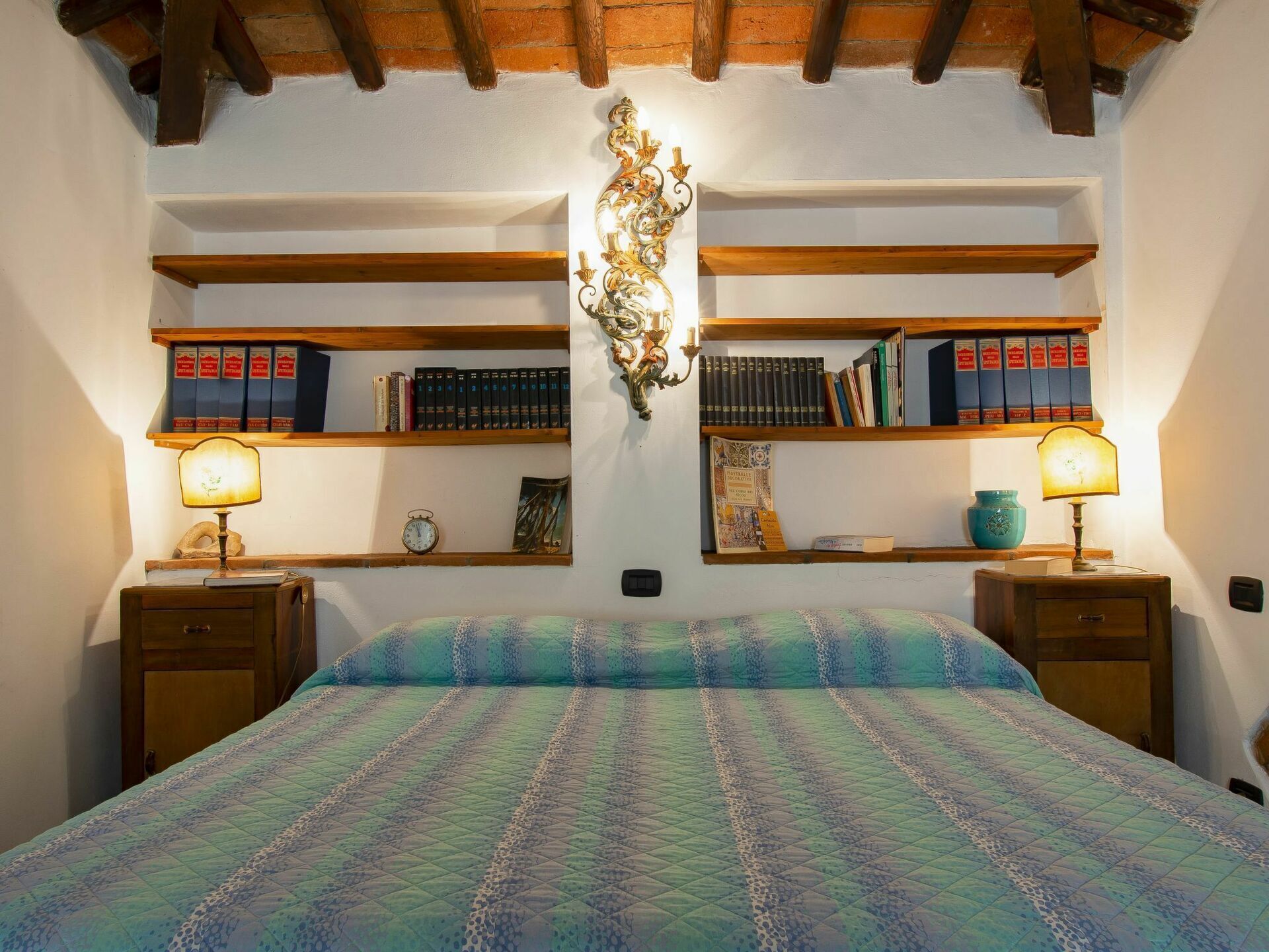 Een slaapkamer met een bed, twee nachtkastjes, een wandplank met boeken, een decoratief wandobject en een klok.