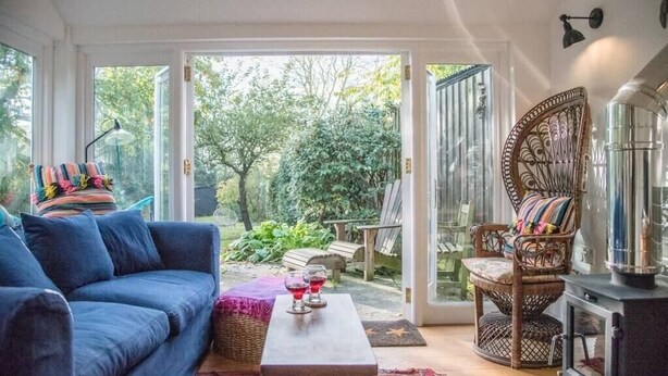 A cozy living room with a blue sofa, wicker chairs, a small table with glasses, and a view of a garden through glass doors. - The Coach House, Orford (Orford)