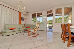 A spacious living room with a sofa, chairs, a dining table, and large windows offering a view of the outdoors.