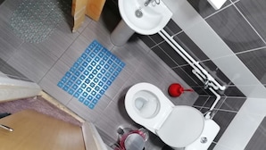 Un baño con inodoro, bidé y lavamanos. Hay una alfombra azul en el suelo y un rociador de bidé rojo.