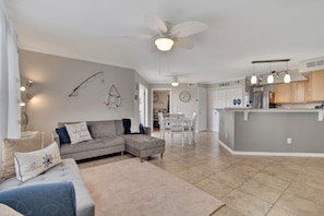 A spacious living room with a grey sectional sofa, a ceiling fan, and a kitchen area with a grey island and wooden cabinets.