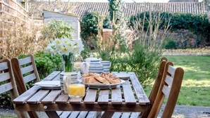 Une table en bois, installée à l’extérieur, est dressée pour le petit-déjeuner : on y trouve des croissants, un verre de jus d’orange et un vase rempli de fleurs blanches.