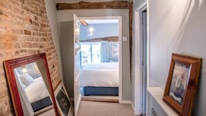 Un couloir étroit avec un mur en briques, une porte en bois et un lit visible à travers une porte ouverte donnant sur la pièce adjacente.