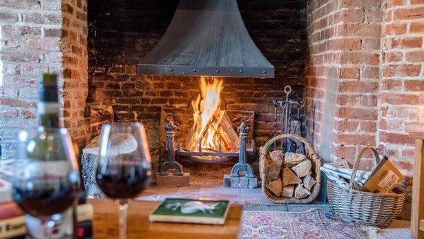 Une pièce chaleureuse avec une cheminée en briques, une table avec des verres à vin et des livres, et un tapis à motifs.