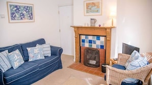 A living room with a blue sofa, a wicker chair, a fireplace with a tiled surround, and two framed artworks on the walls.
