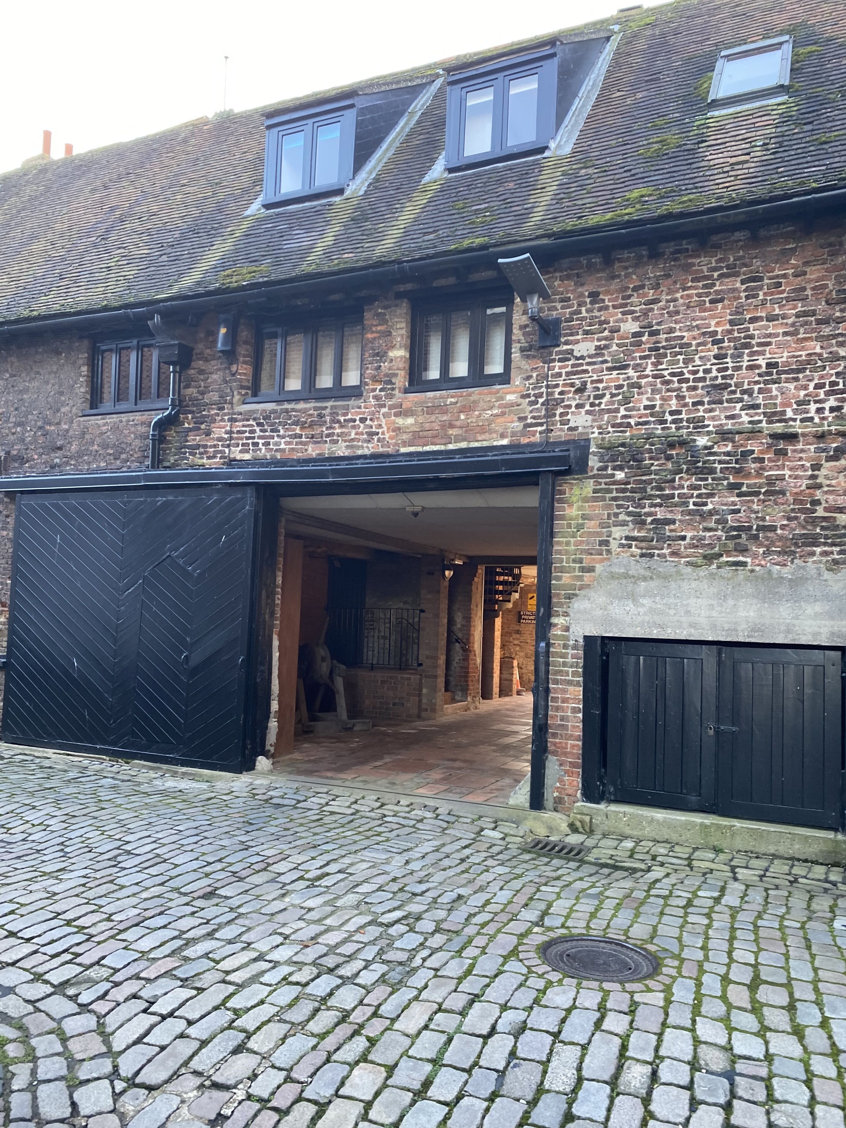 A traditional brick building with a large garage door, a cobblestone driveway, and a sloped roof with dormer windows.