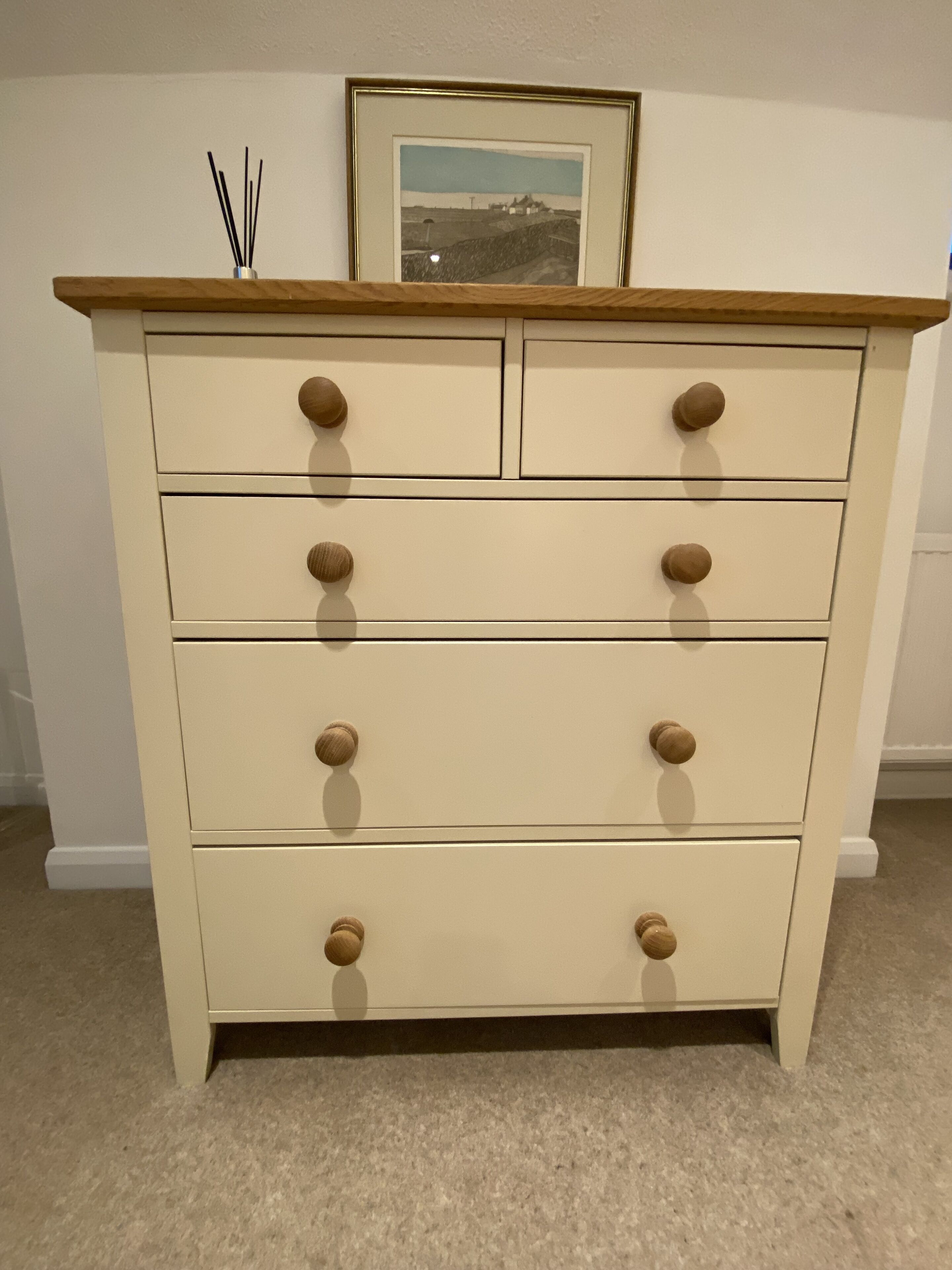 A cream-colored dresser with wooden knobs, a framed picture above it, and a small vase with reeds on the top.