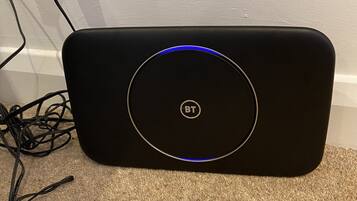 A black speaker with a circular logo and a blue light on the front, connected to a power strip with a white plug.