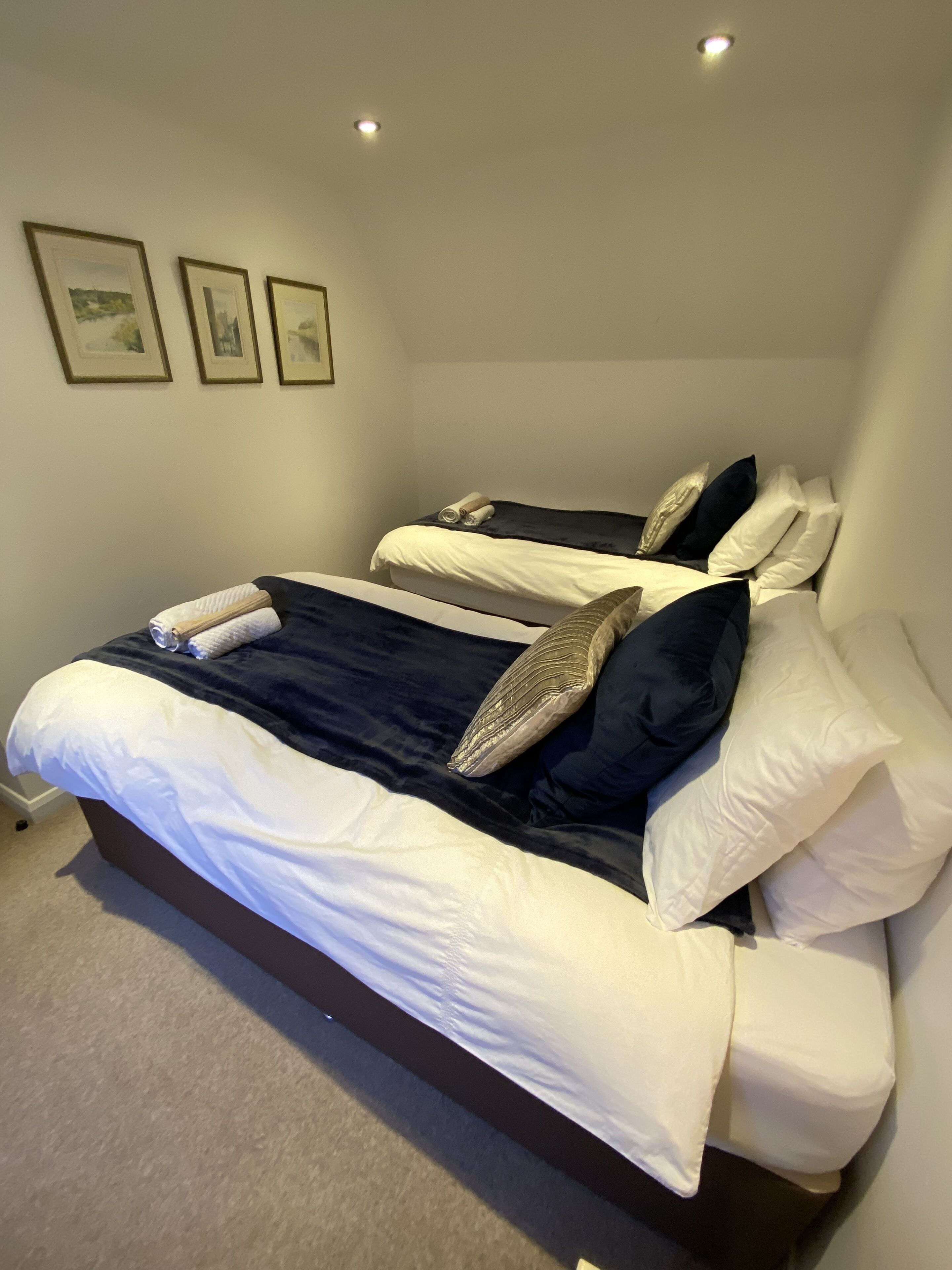 Un dormitorio con dos camas, cada una con sábanas blancas y ropa de cama azul oscuro. Hay tres cuadros en la pared.