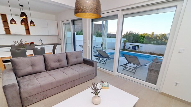 Una sala de estar moderna con un sofá gris, una mesa de centro blanca y vista a una piscina exterior a través de grandes puertas correderas de vidrio.
