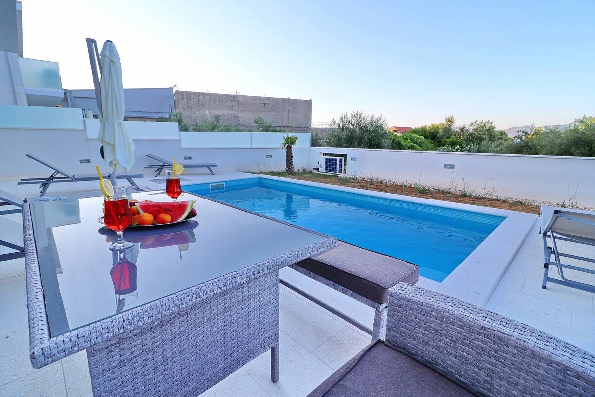 A modern outdoor pool area with a glass table, wicker chairs, and a fruit bowl with a drink.