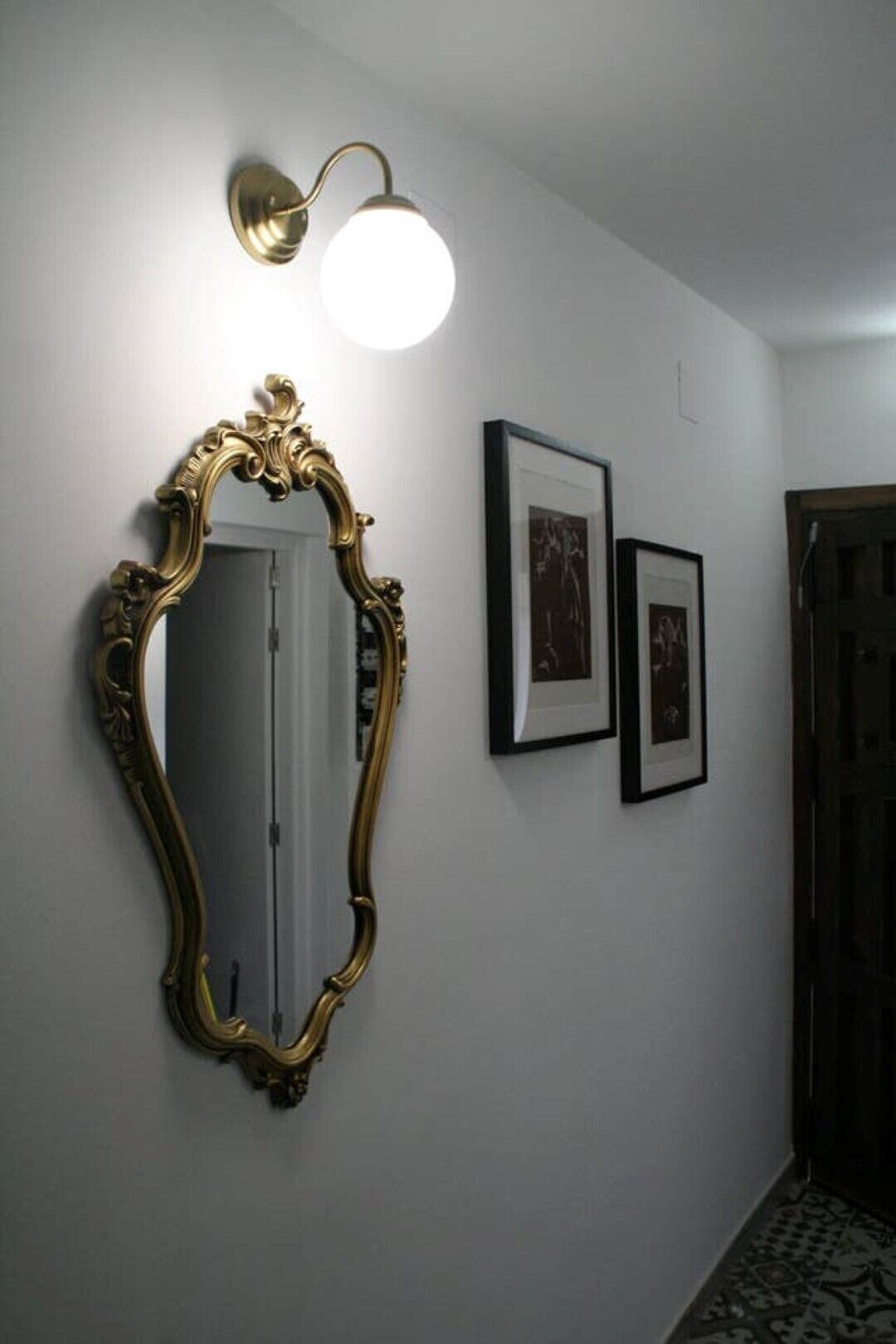 Un pasillo con un espejo grande y ornamentado, una lámpara de pared y dos cuadros enmarcados.