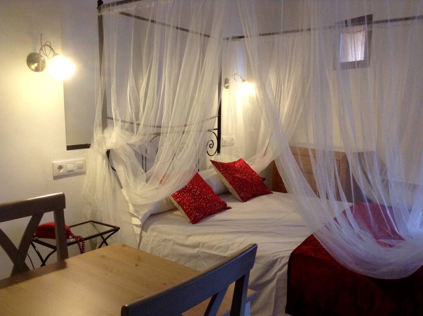 Une pièce avec un lit recouvert d’un voile blanc, deux oreillers rouges, une table en bois et une chaise.