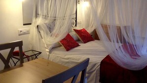 Ein Zimmer mit einem Bett, das mit weißem Netz verhängt ist, zwei roten Kissen, einem Holztisch und einem Stuhl.