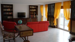 A living room with a red sofa, a wooden coffee table, a globe, and a television. There are bookshelves and a window with yellow curtains.